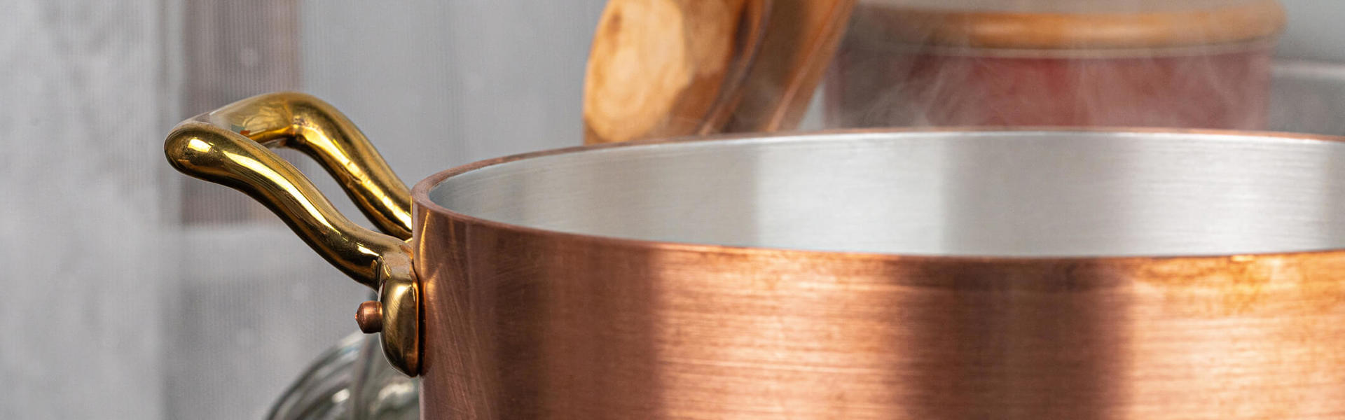 a close up of a copper cooking pan with brass handles and wooden cooking utensils in the background