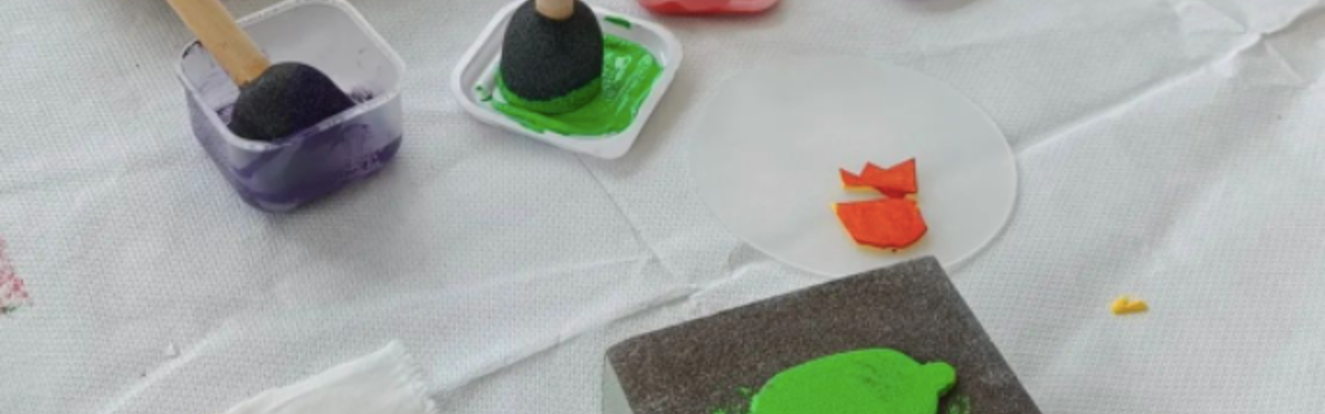 Homemade stamp making tools on a white table cloth in various primary colour paints