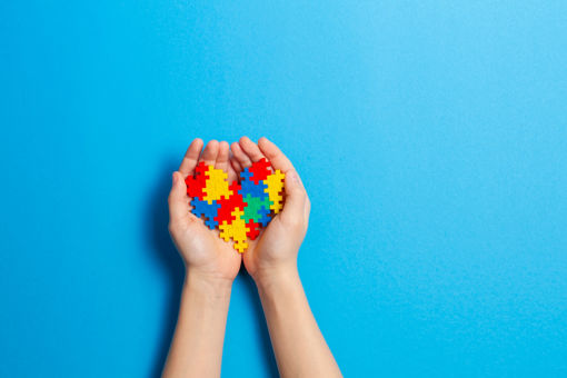 Hands holding a multicoloured jigsaw heart, representing Autism Acceptance Month, against a blue background.