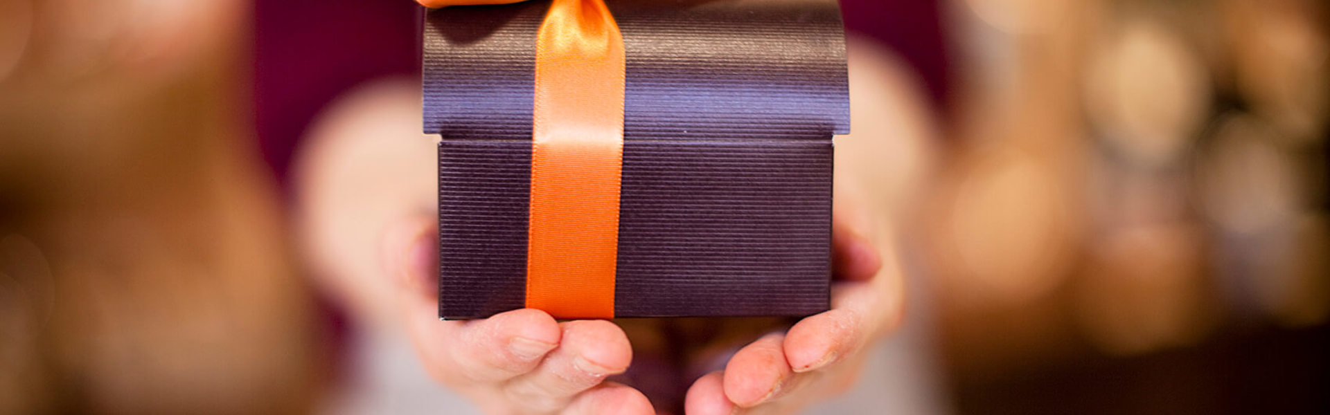 a pair of hands holding a a satin black gift box wrapped in an orange ribbon