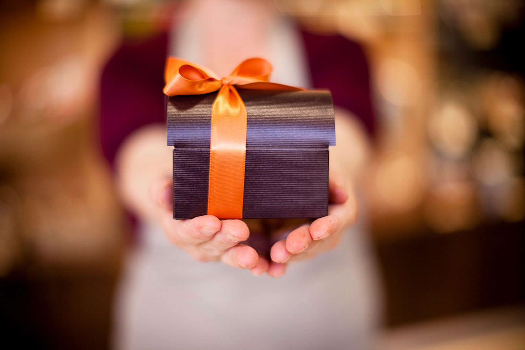 a pair of hands holding a a satin black gift box wrapped in an orange ribbon