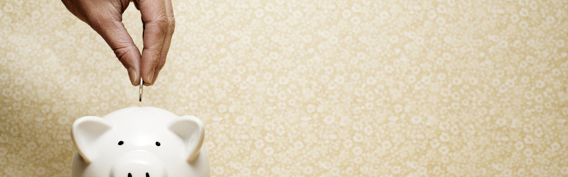 a hand placing a penny into a white piggy bank against a yellow floral wallpaper background