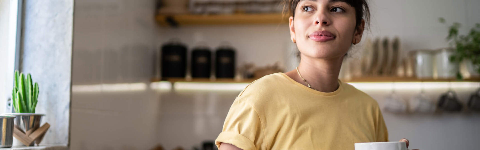 a woman in a yellow t-shirt holding a mug of tea looking out of her window