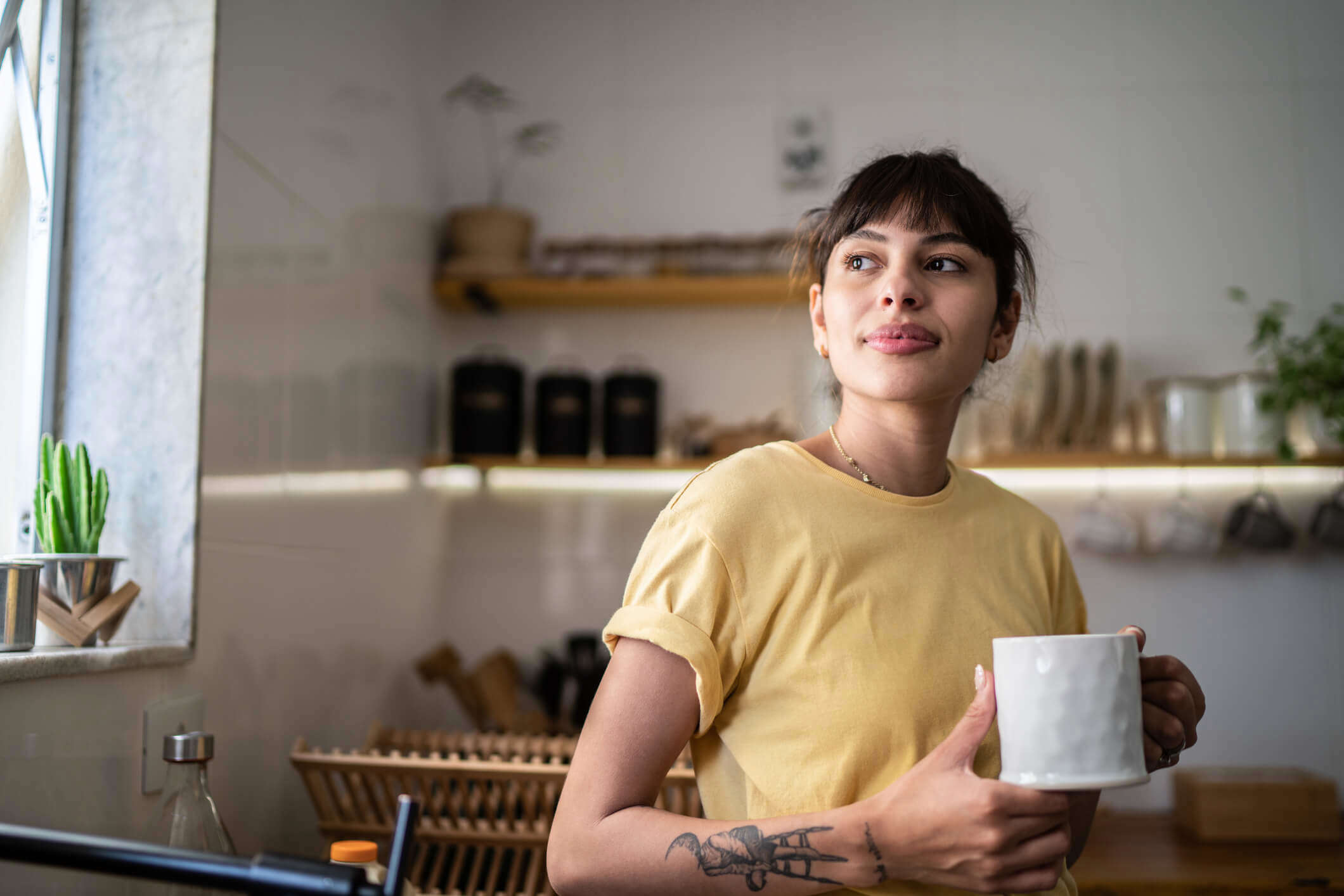 a woman in a yellow t-shirt holding a mug of tea looking out of her window