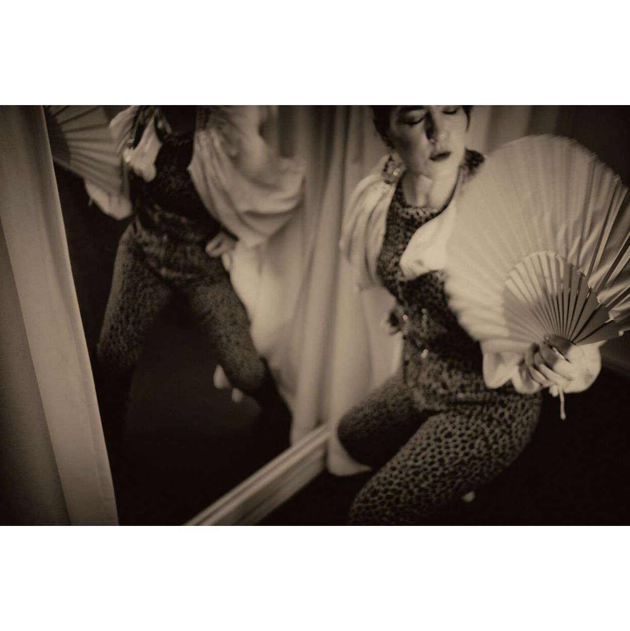 A black and white photo of a woman in a leopard print bodysuit practising burlesque poses in the mirror holding a hand fan.