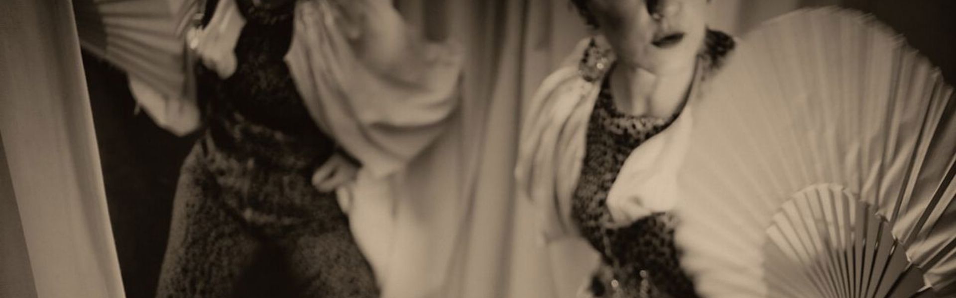 A black and white photo of a woman in a leopard print bodysuit practising burlesque poses in the mirror holding a hand fan.