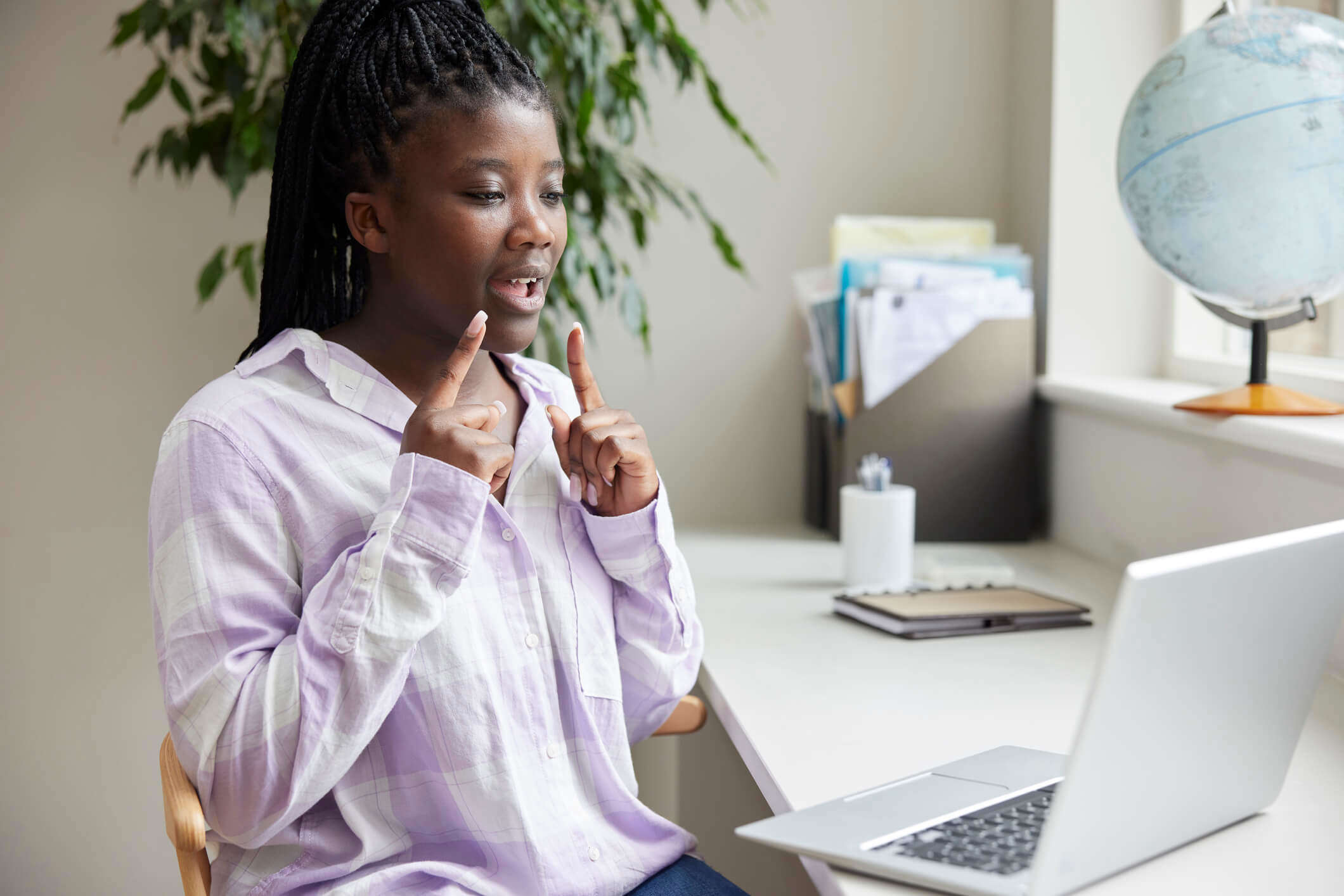 a woman wearing a purple and white check shirt practicing sign language using her laptop at a desk