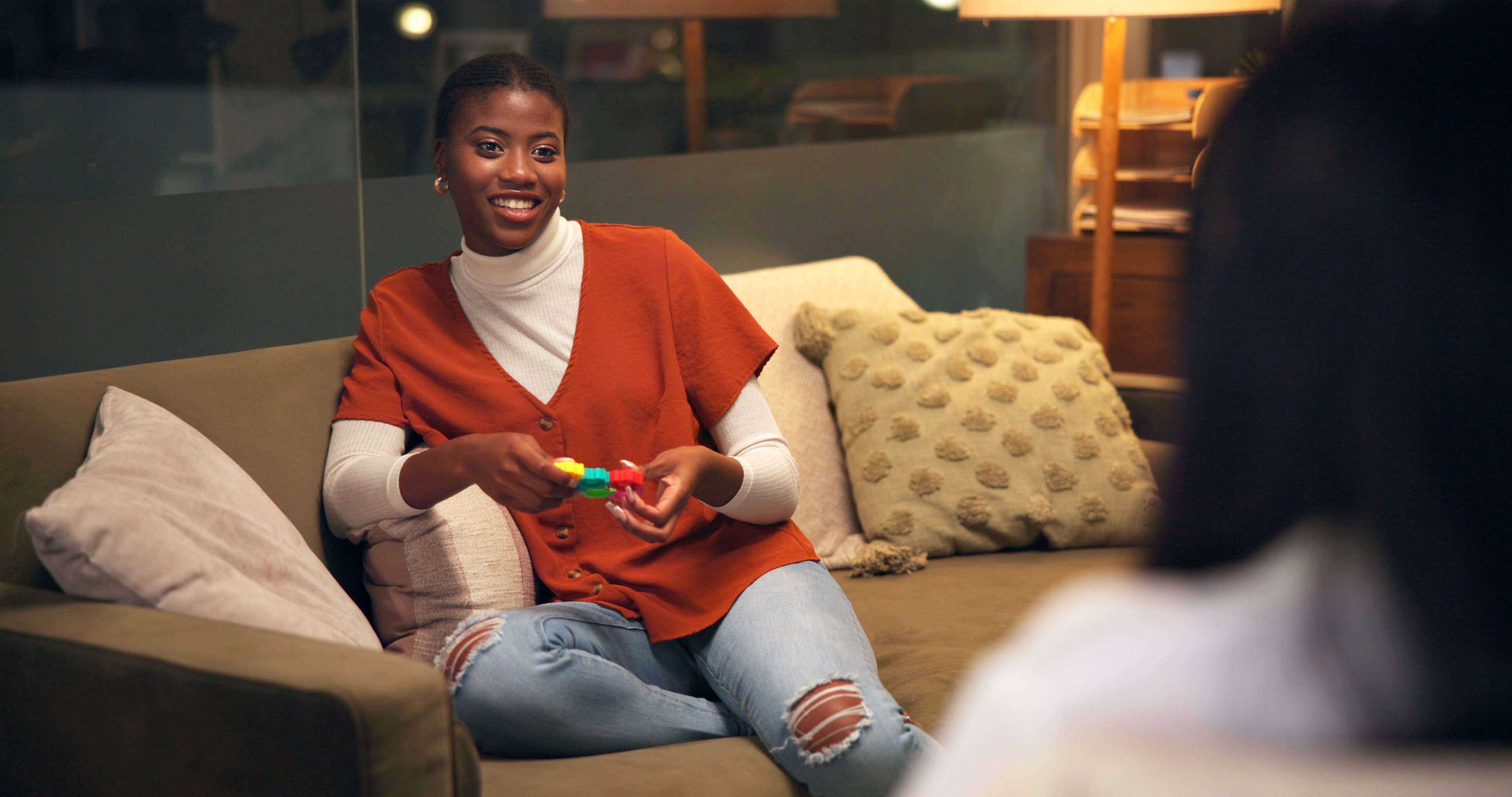 Main image: a woman sat on a couch smiling, stimming with a fidget toy.