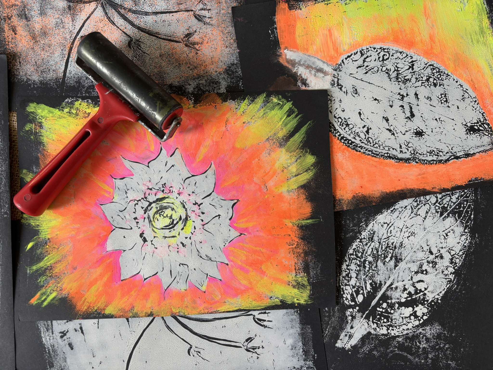 Main Image: A selection of monoprints, on dark paper with neon orange, yellow, pink and silver paints, with a red printing roller.