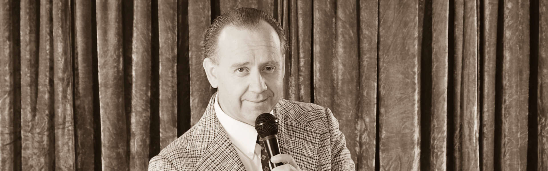 A sepia toned photo of quiz master David Mustoe holding a microphone in one hand and a card with question marks in the other