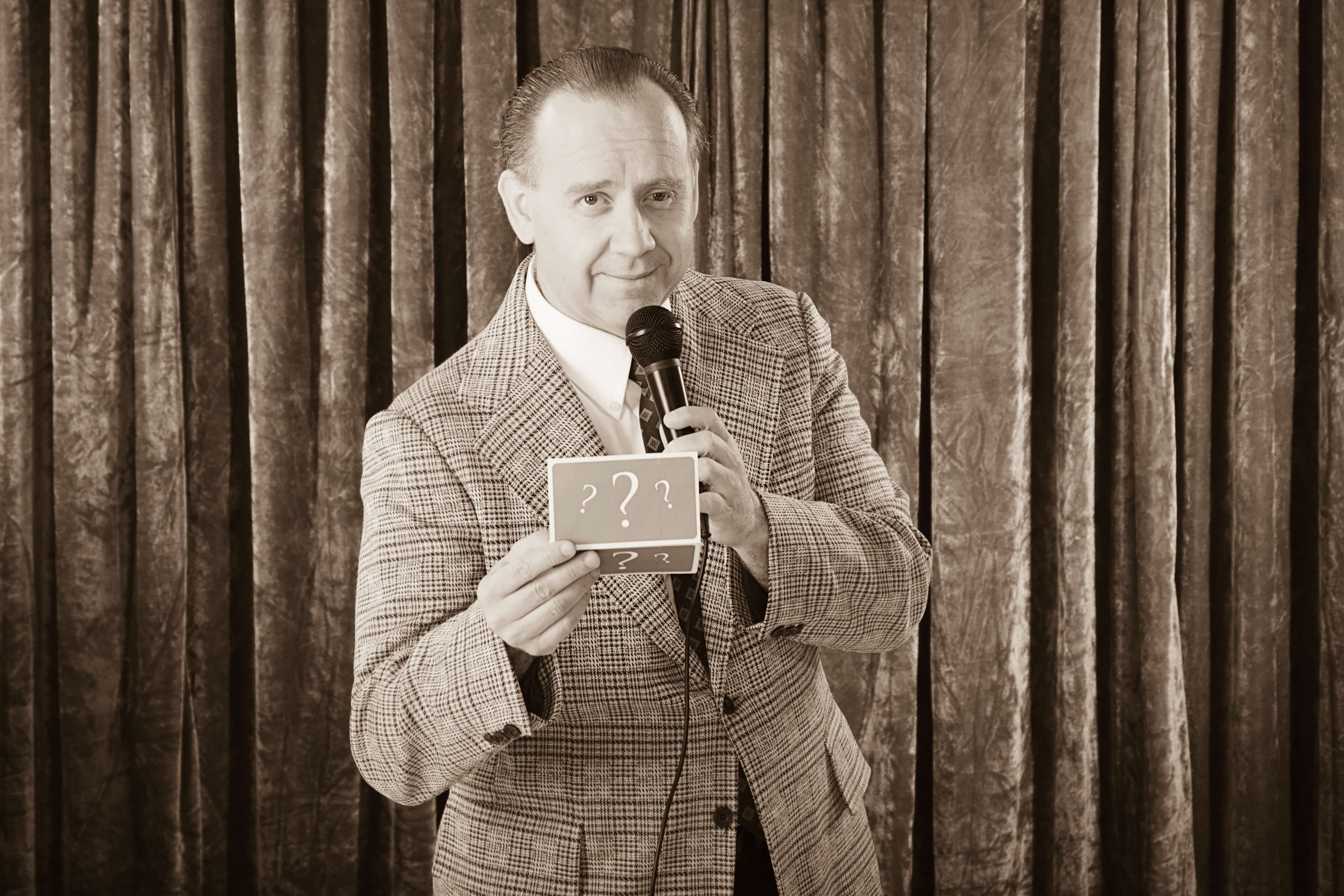 A sepia toned photo of quiz master David Mustoe holding a microphone in one hand and a card with question marks in the other