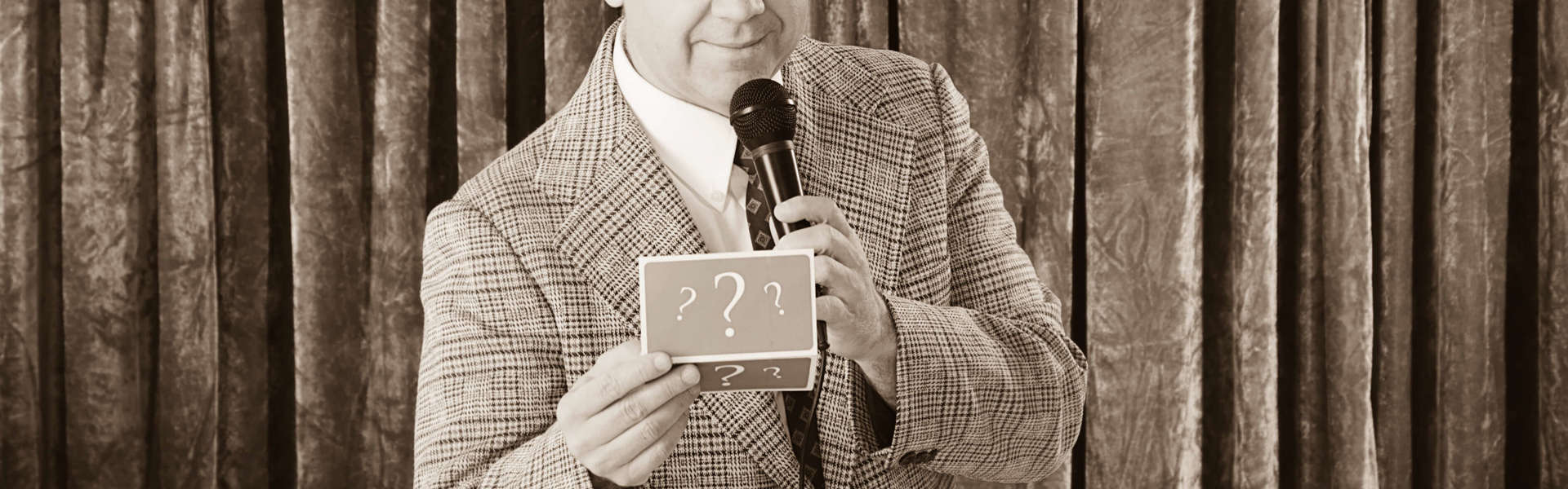 A sepia toned photo of quiz master David Mustoe holding a microphone in one hand and a card with question marks in the other