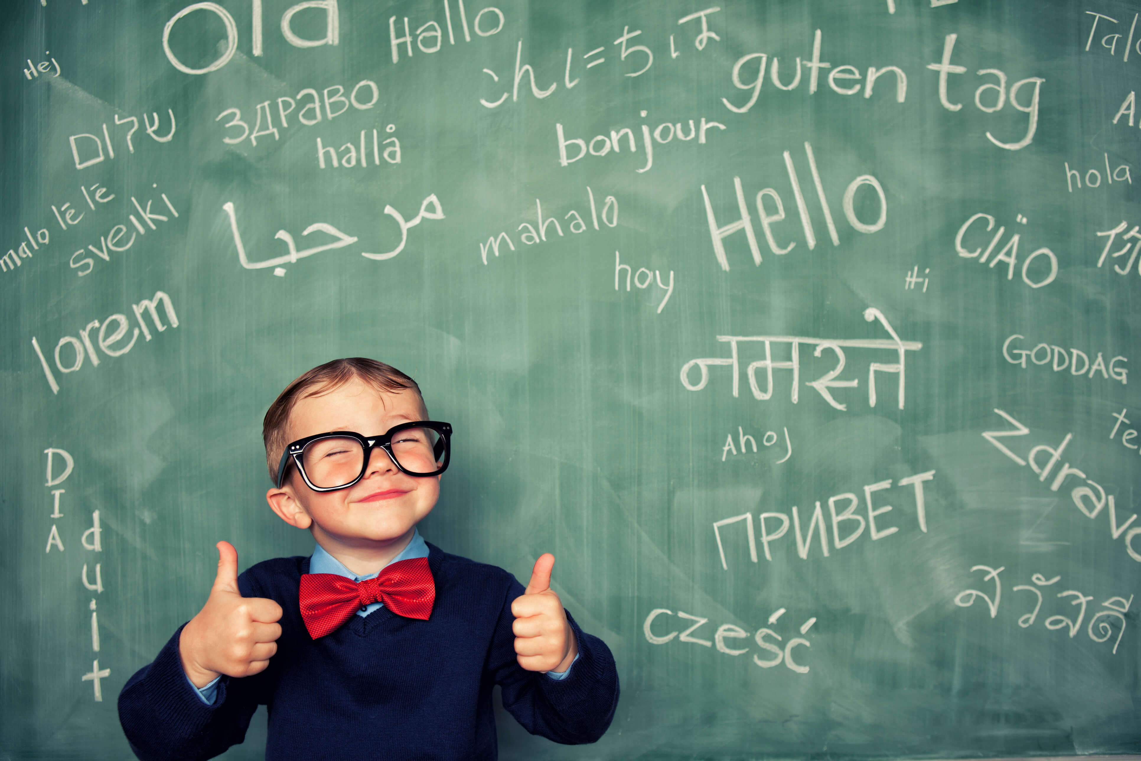 A young boy standing in front of a blackboard displaying 'hello' written in different languages