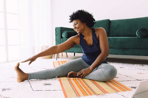 a woman sat on the living room floor on a yoga mat doing an online yoga class on her laptop