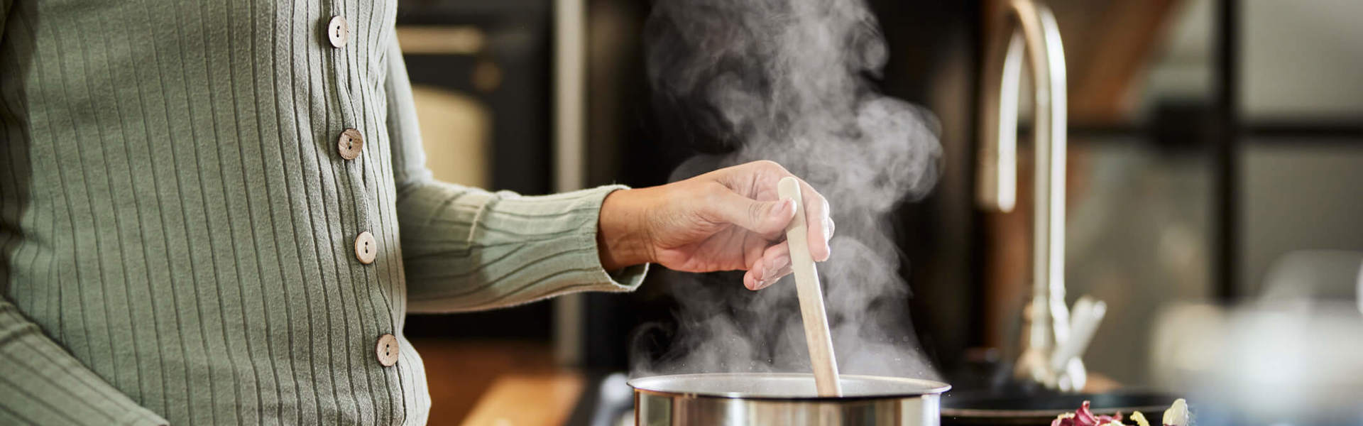 a woman wearing a green jumper stirring a boiling soup with a wooden spoon in a silver pan