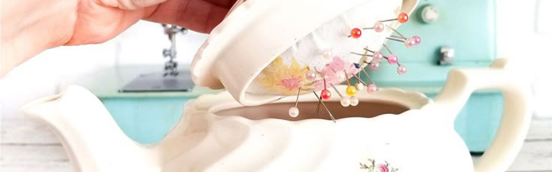 DIY Teapot Sewing Kit - a while teapot with a floral design on the front, with the lid being opened by a hand to reveal a pincushion inside the lid