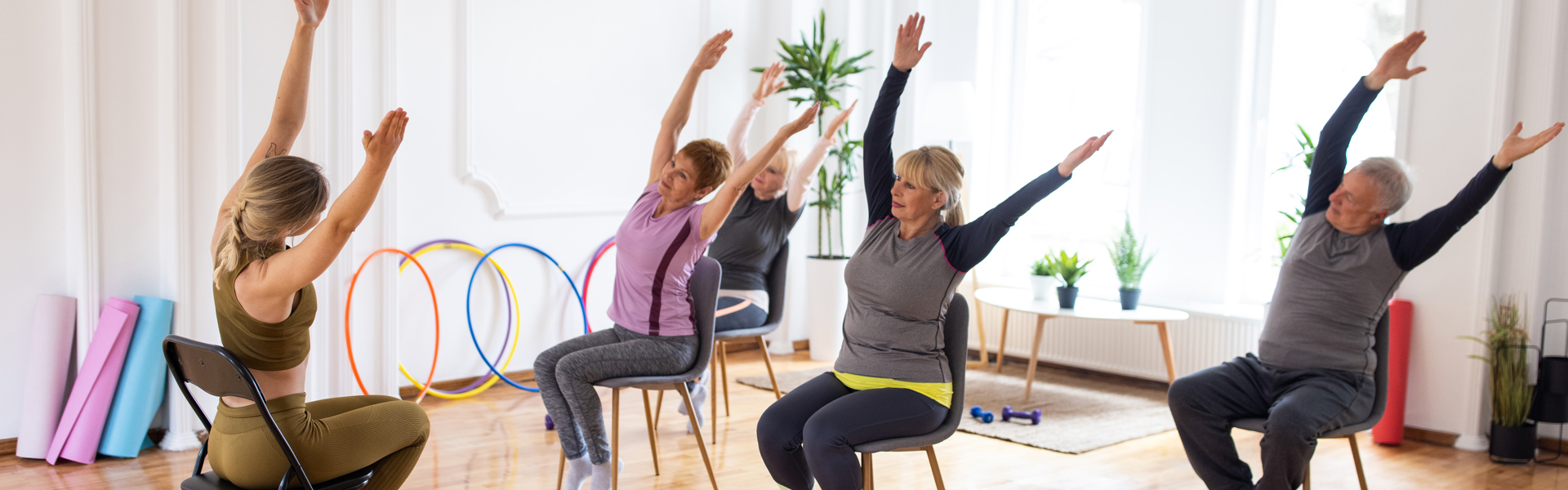 Main Image: a group of people, sat on chairs doing seated pilates, with an instructor demonstrating at the front.