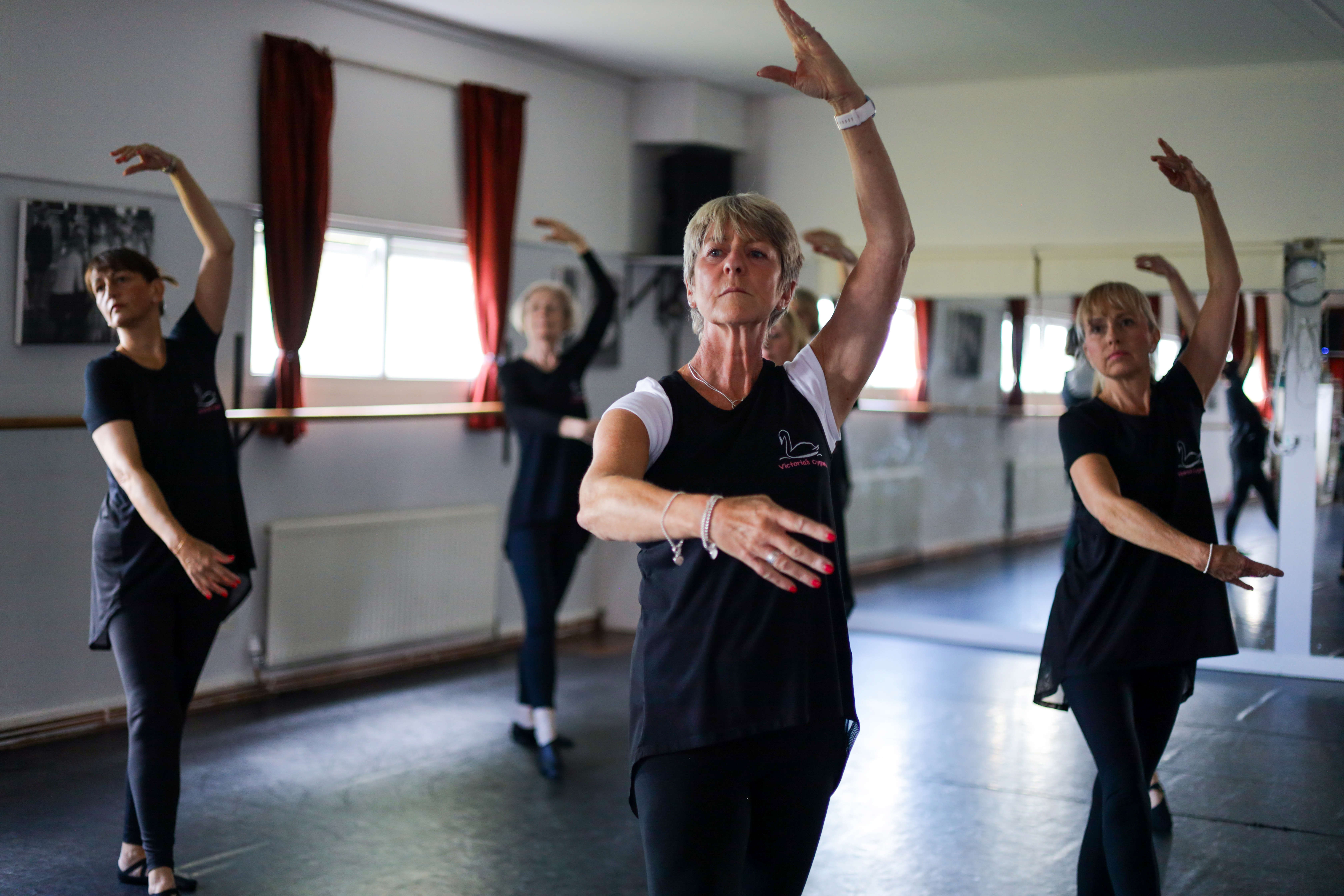 A group of four middle-aged women, all wearing black, holding a ballet pose, with one arm out in front of their bodies and one raised above their heads, with serious expressions on their face.