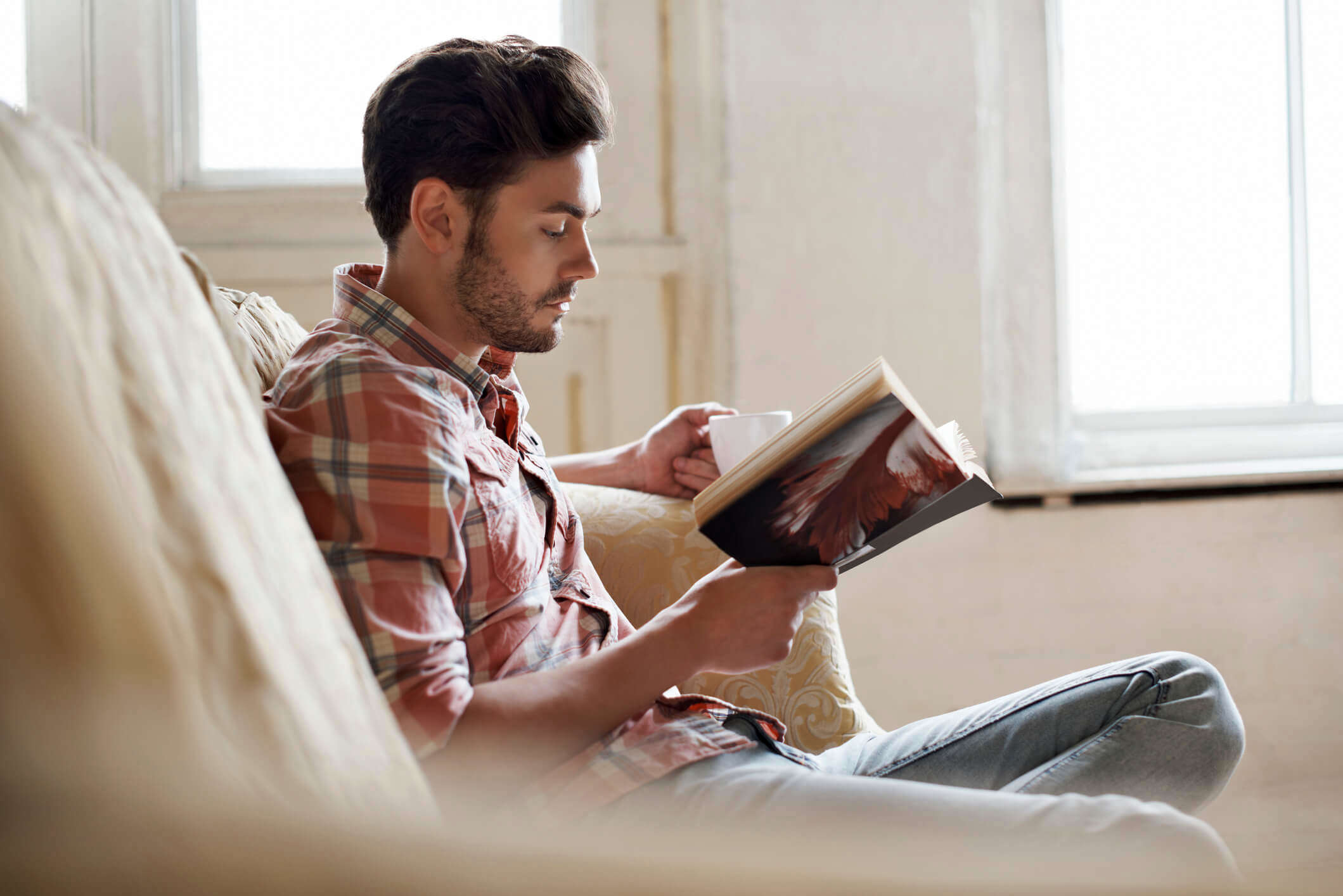 a man sat on the sofa with a cup of tea reading a book