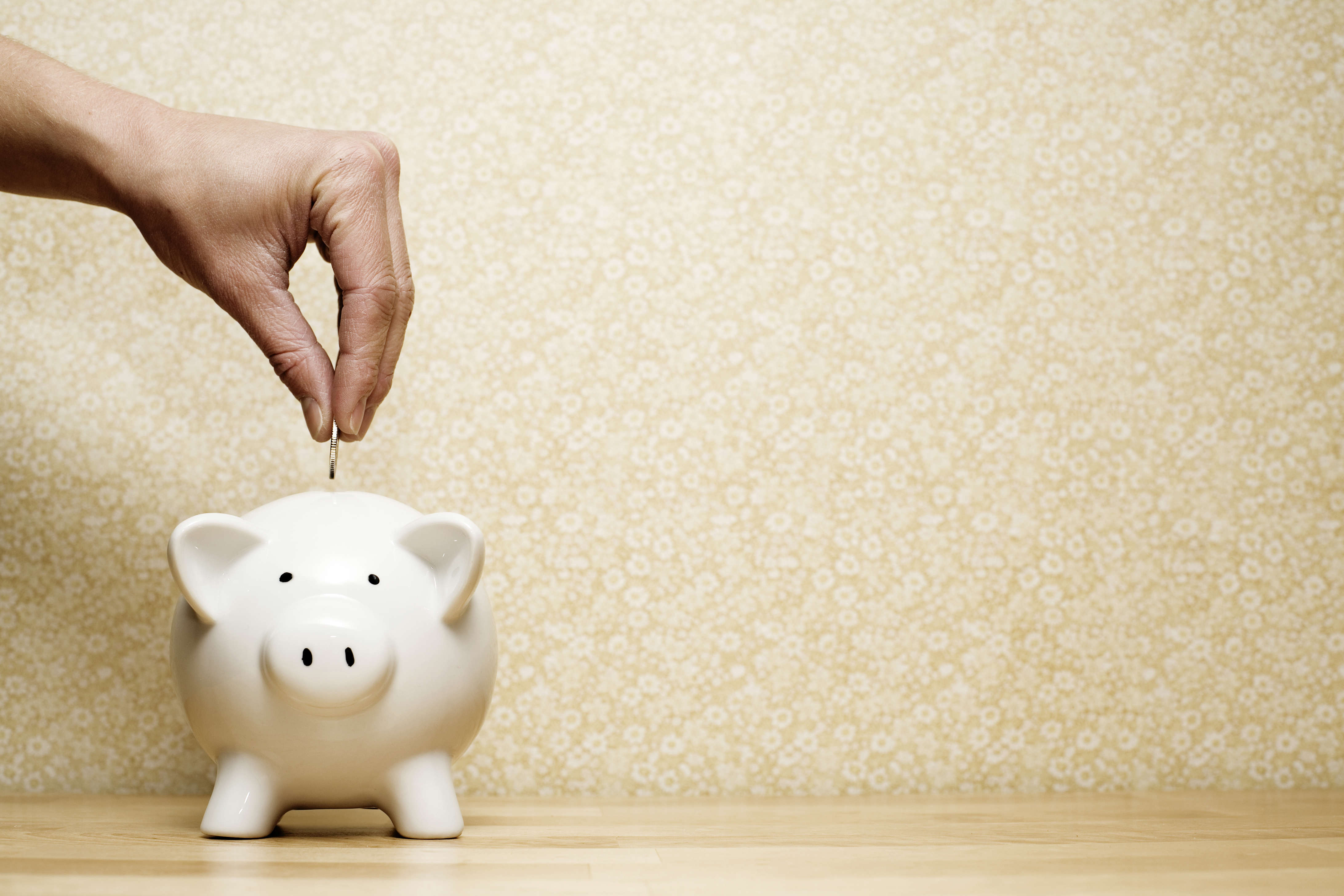 a hand putting a silver coin into a white piggy bank money box, resting on a wooden table with a yellow wallpaper background