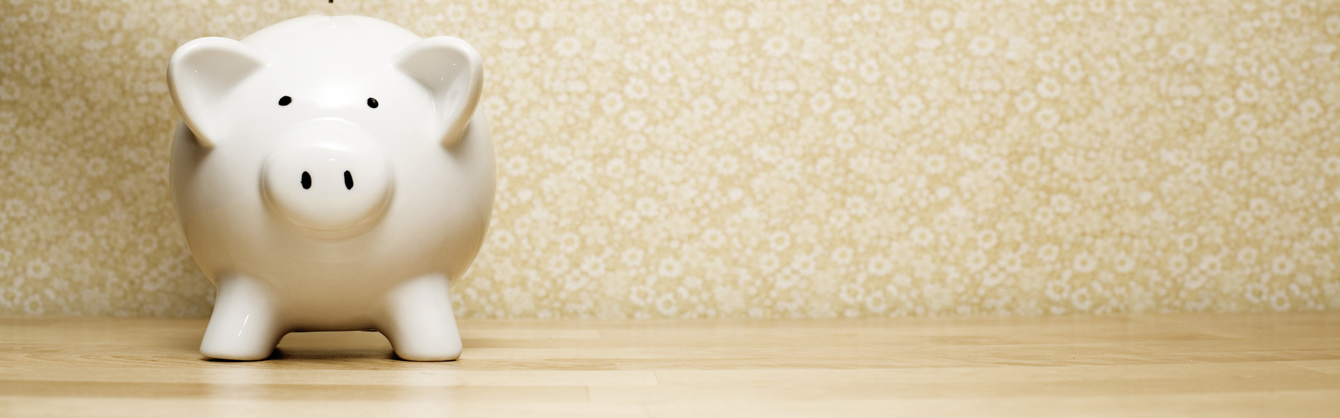 a hand putting a silver coin into a white piggy bank money box, resting on a wooden table with a yellow wallpaper background