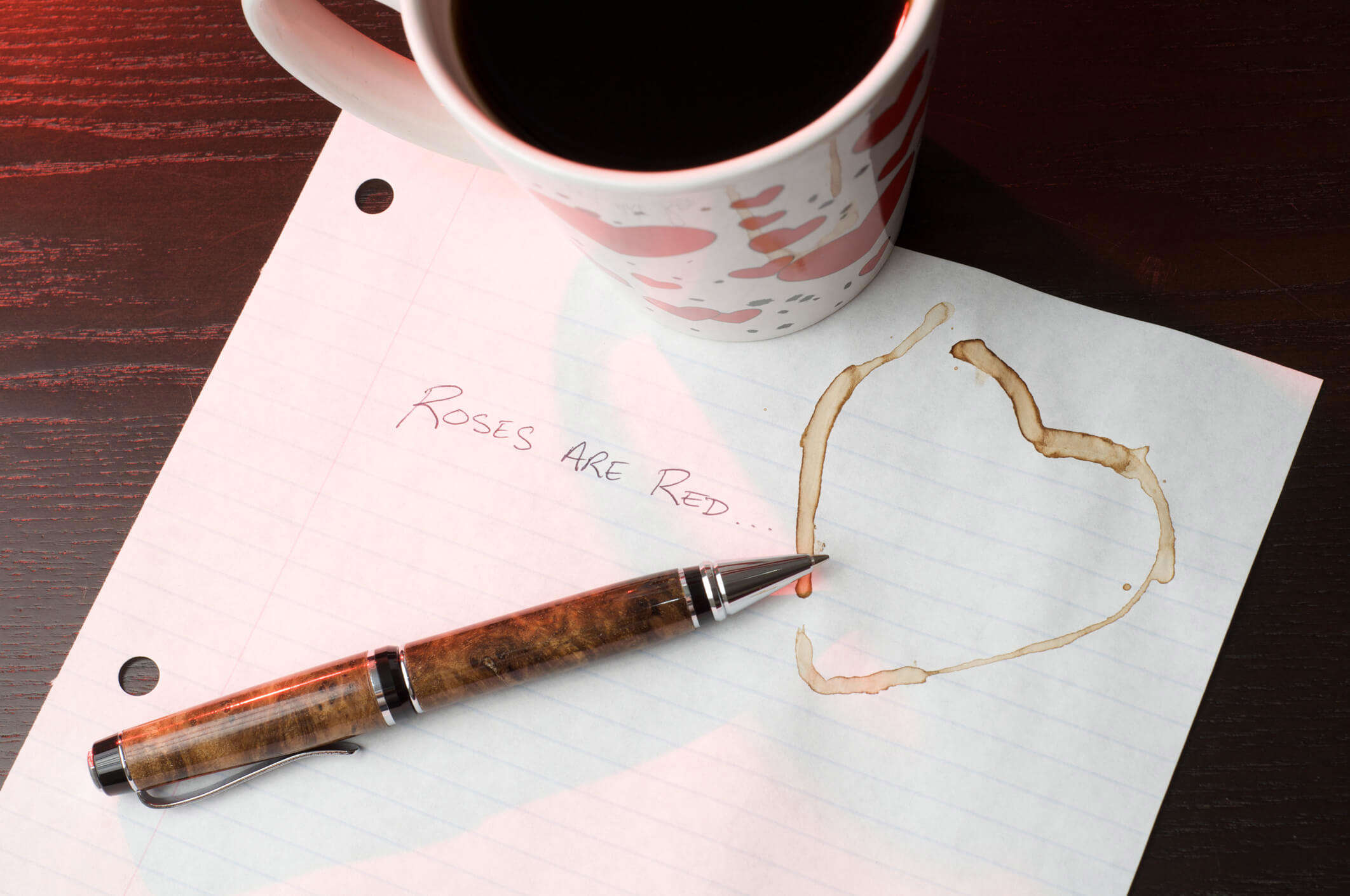 a mug of coffee resting on a valentine's day poem next to a pen and heart shaped coffee mark