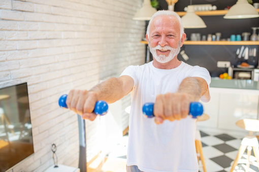An older man wearing a white t-shirt holding small blue weights in front of him in his kitchen