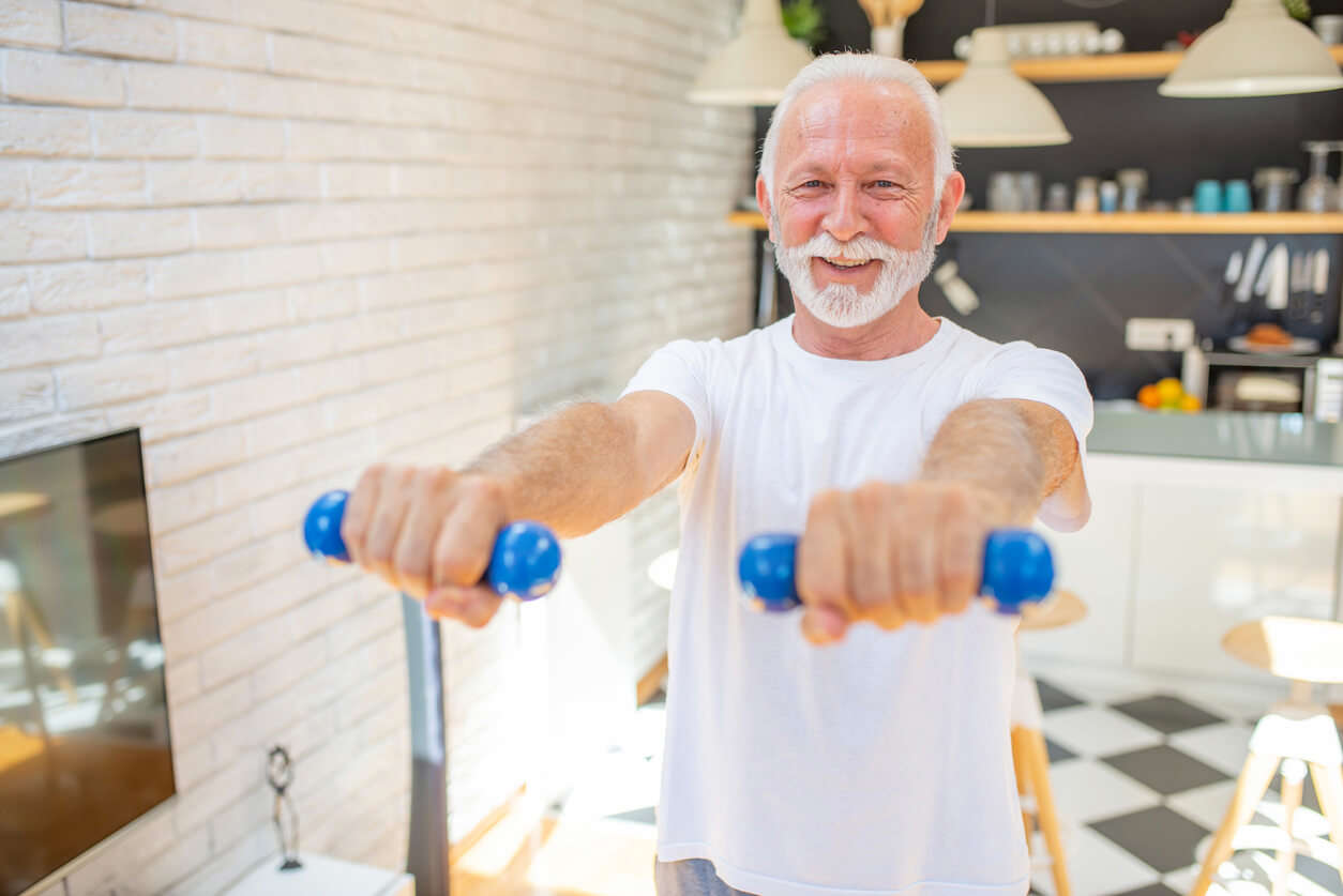 An older man wearing a white t-shirt holding small blue weights in front of him in his kitchen