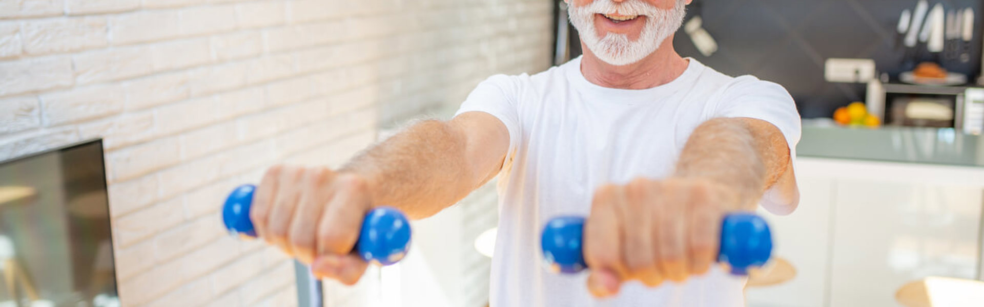 An older man wearing a white t-shirt holding small blue weights in front of him in his kitchen