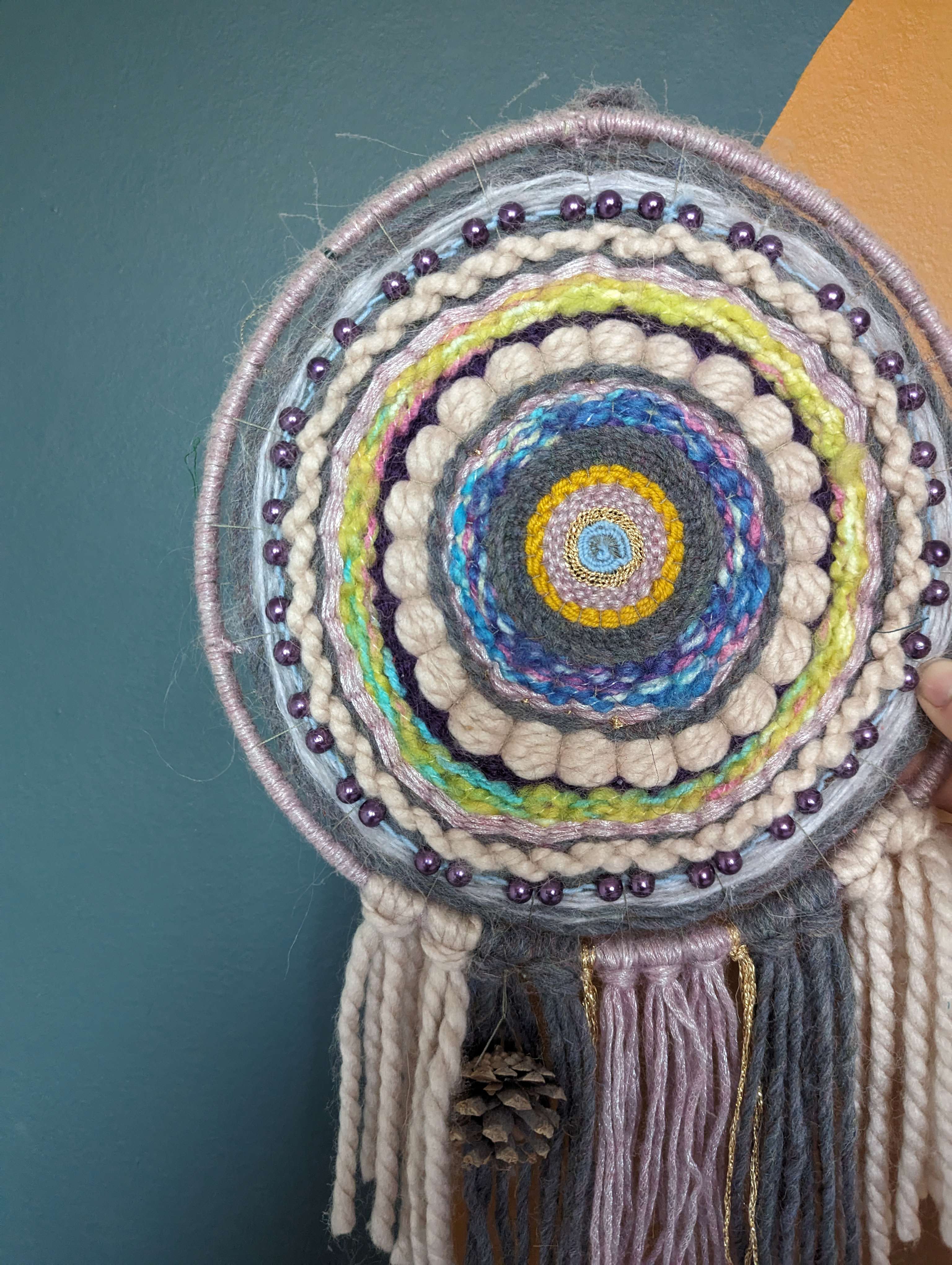 multi-coloured weaved wall hanging in a variety of string and thread material against a teal blue background