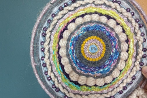 multi-coloured weaved wall hanging in a variety of string and thread material against a teal blue background