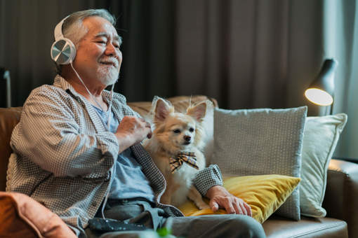 A person sitting on the sofa with their Chihuahua dog, listening to music through headphones, smiling.