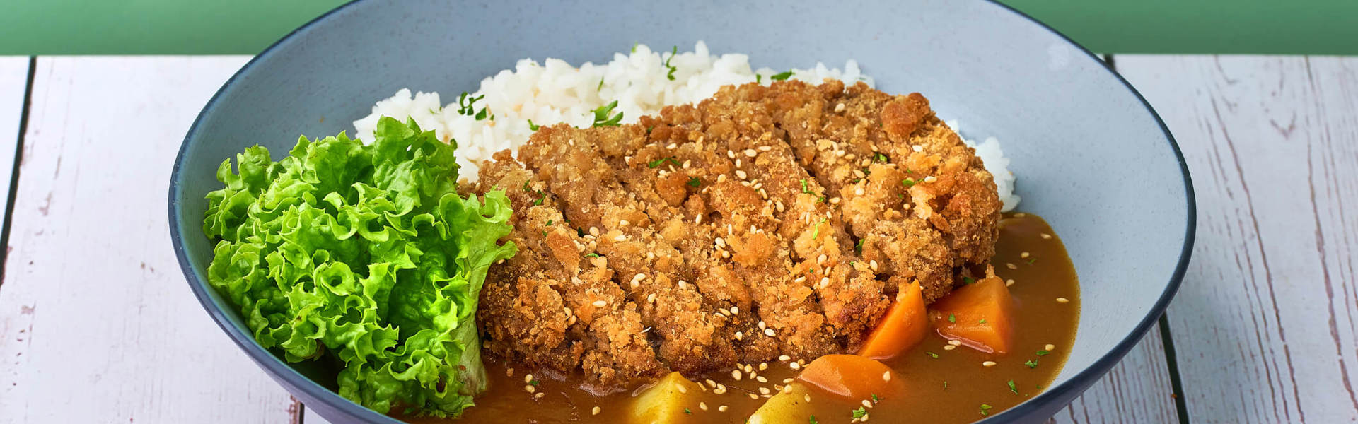 Chicken katsu curry with white rice and leafy greens in a blue bowl on a green background