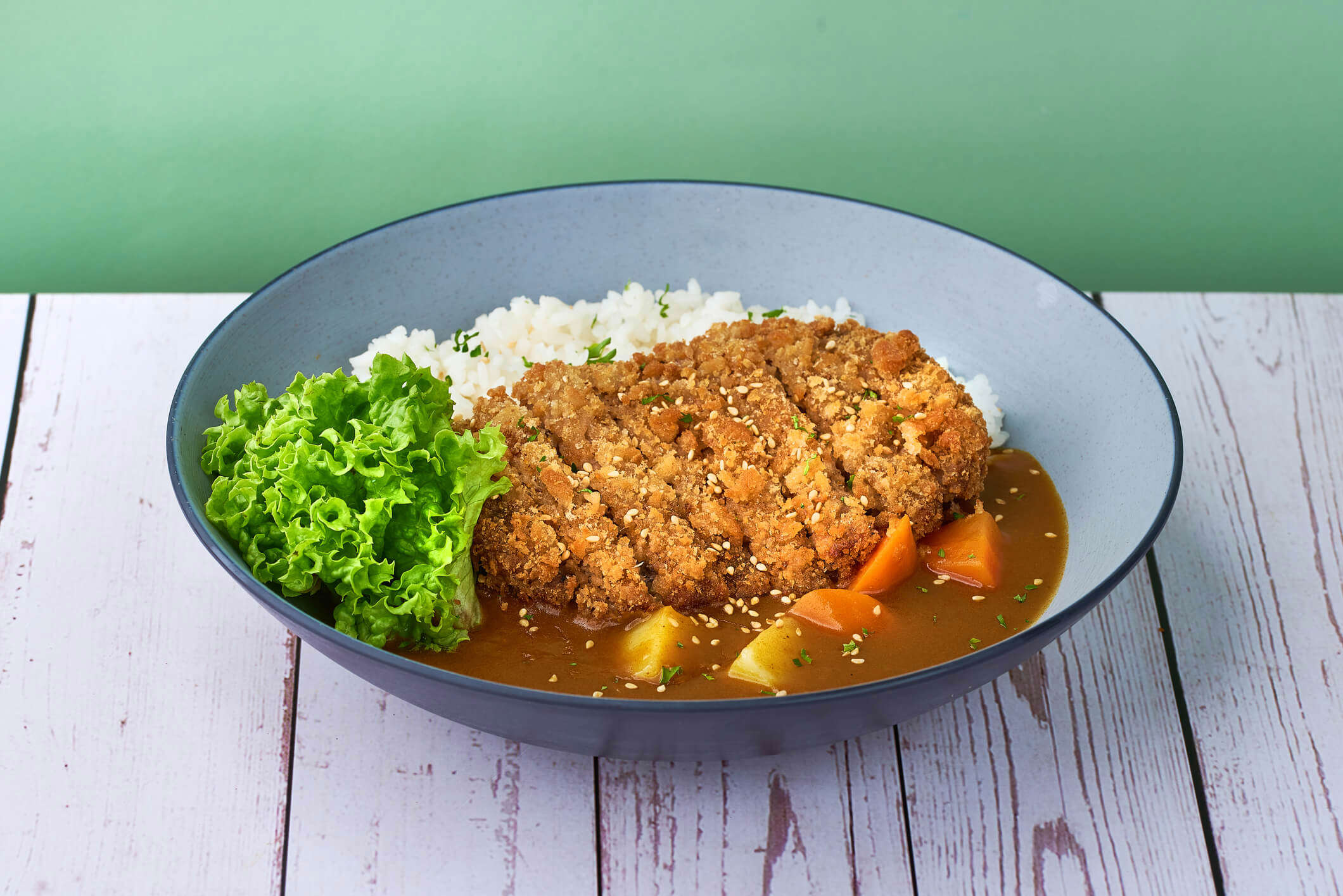 Chicken katsu curry with white rice and leafy greens in a blue bowl on a green background