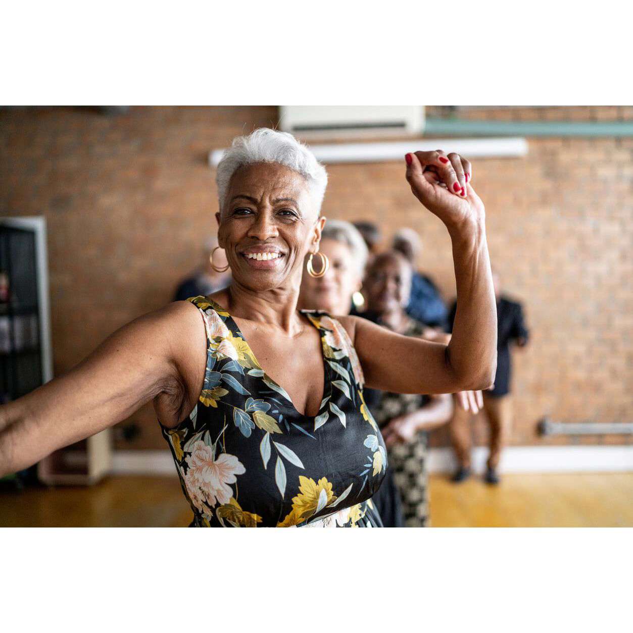 Woman dancing with one arm in the air, smiling wearing a black dress with a yellow and blue floral pattern.