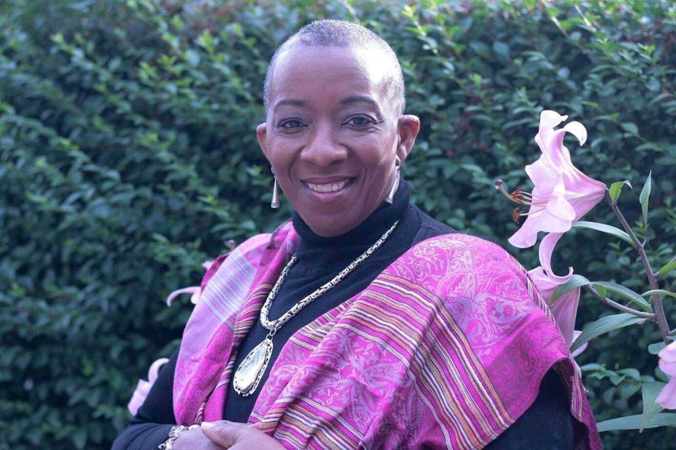 Main Image: a photo of Shirley May, Black British poet, smiling at the camera outside, wearing pink and black, beside a pink flower in the garden.