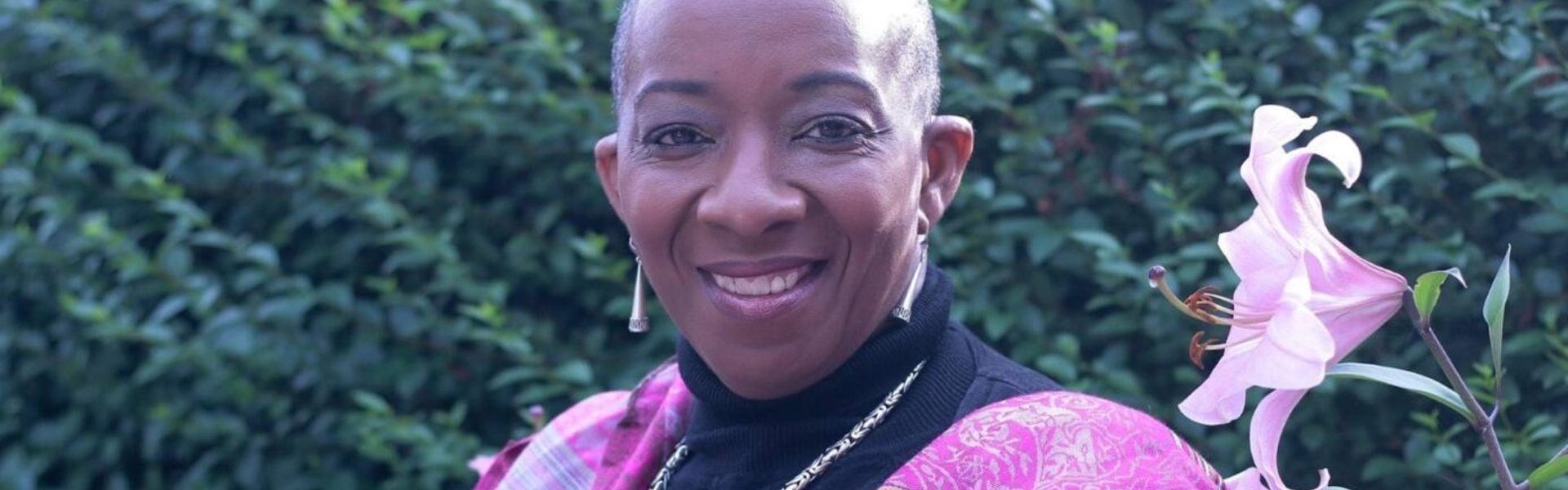 Main Image: a photo of Shirley May, Black British poet, smiling at the camera outside, wearing pink and black, beside a pink flower in the garden.