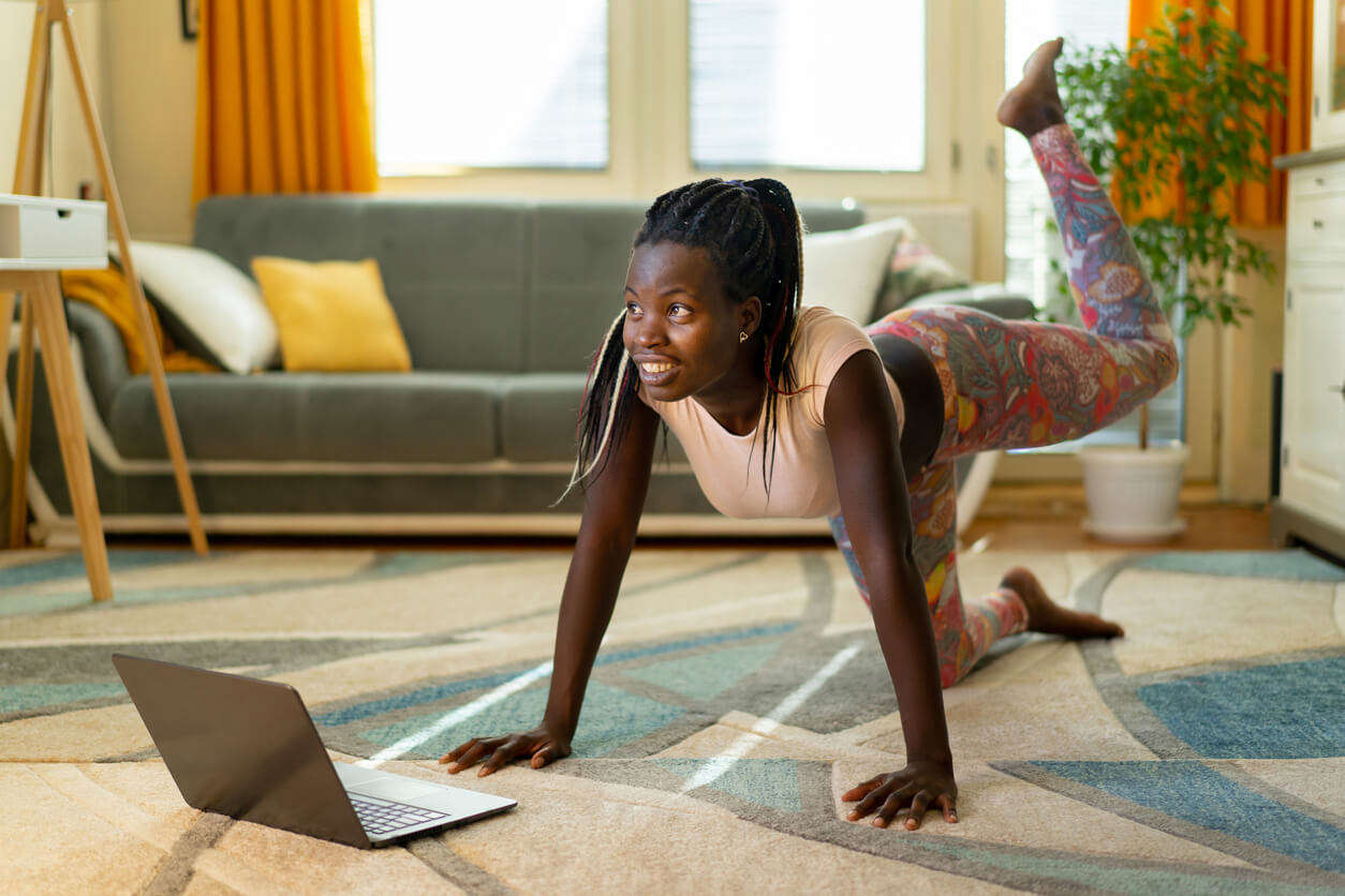 An adult woman does yoga and strength training exercises on a rug in her living room. She is following an online exercise course video on her laptop.