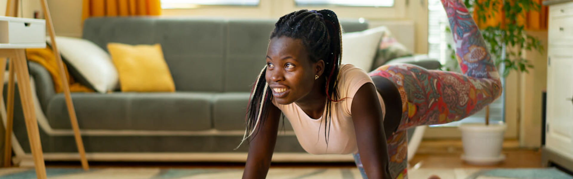 An adult woman does yoga and strength training exercises on a rug in her living room. She is following an online exercise course video on her laptop.