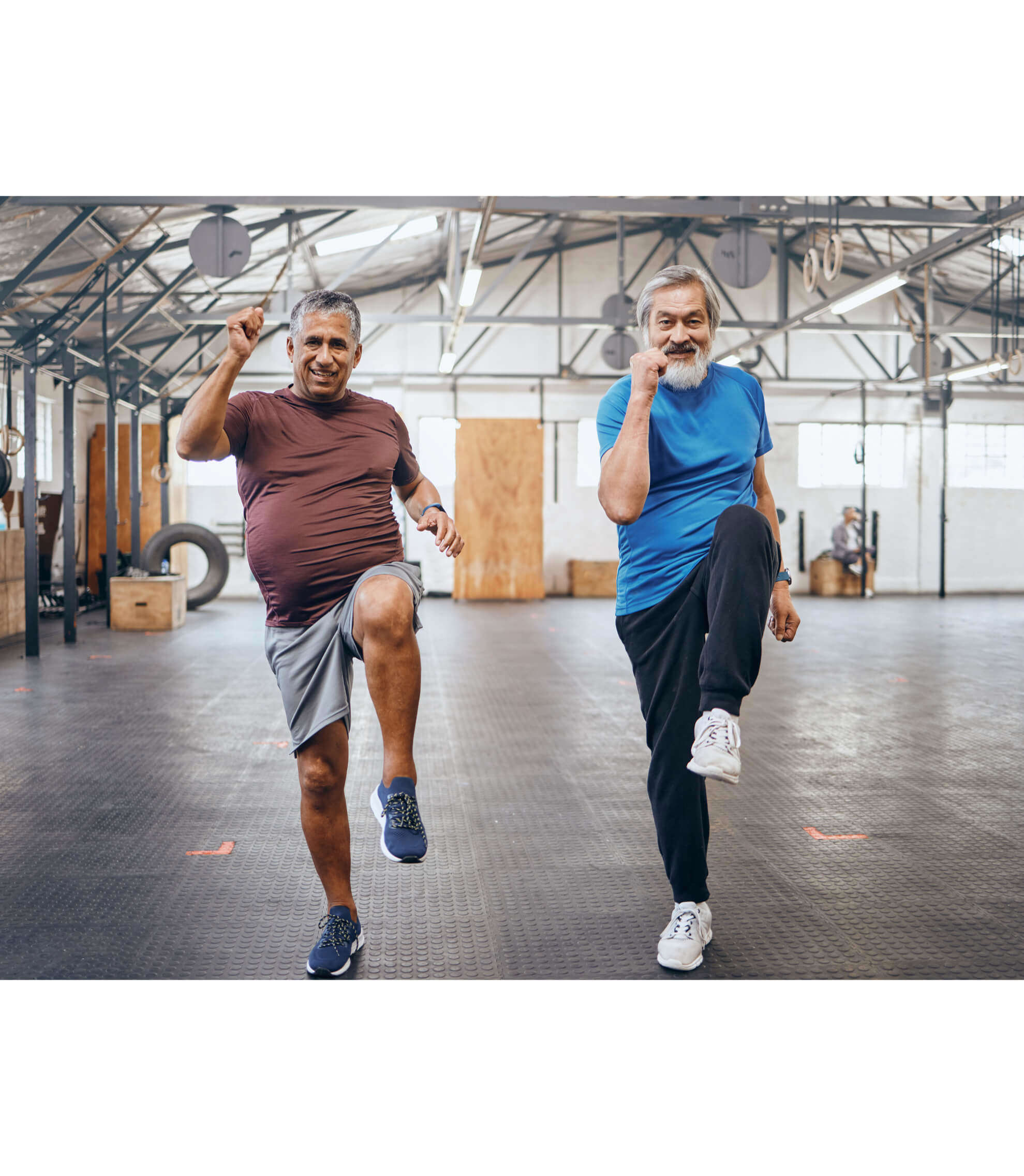 Two older men marching on the spot, enjoying a high intensity workout, smiling, with an exercise hall backdrop.