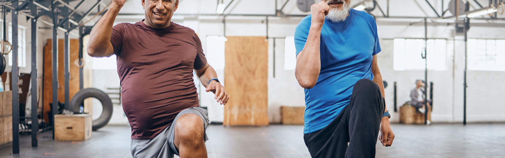 Two older men marching on the spot, enjoying a high intensity workout, smiling, with an exercise hall backdrop.