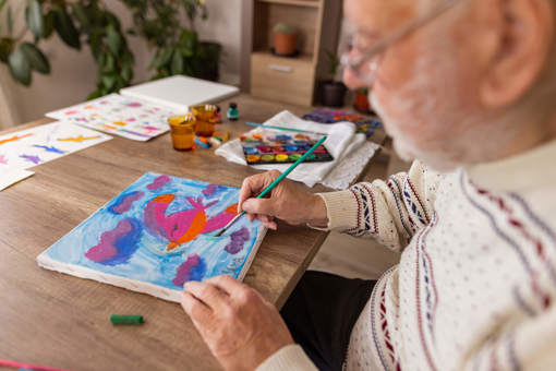A man sat at a table with paintings, paints and a water cup - painting a pink and orange bird, flying though purple and blue clouds.