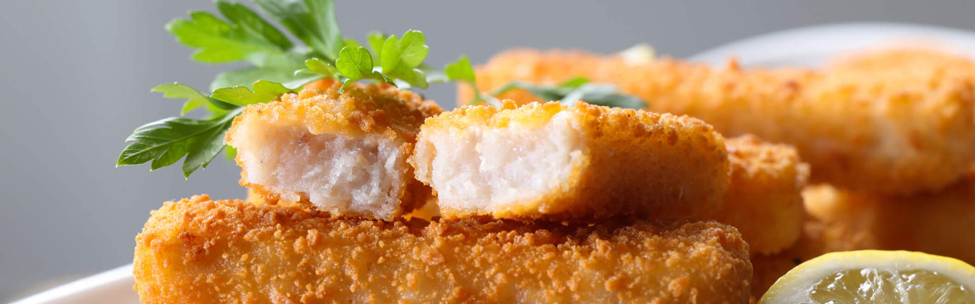 a close up of a pile of homemade fish fingers on a white plat with a slice of lemon and garnished with herbs