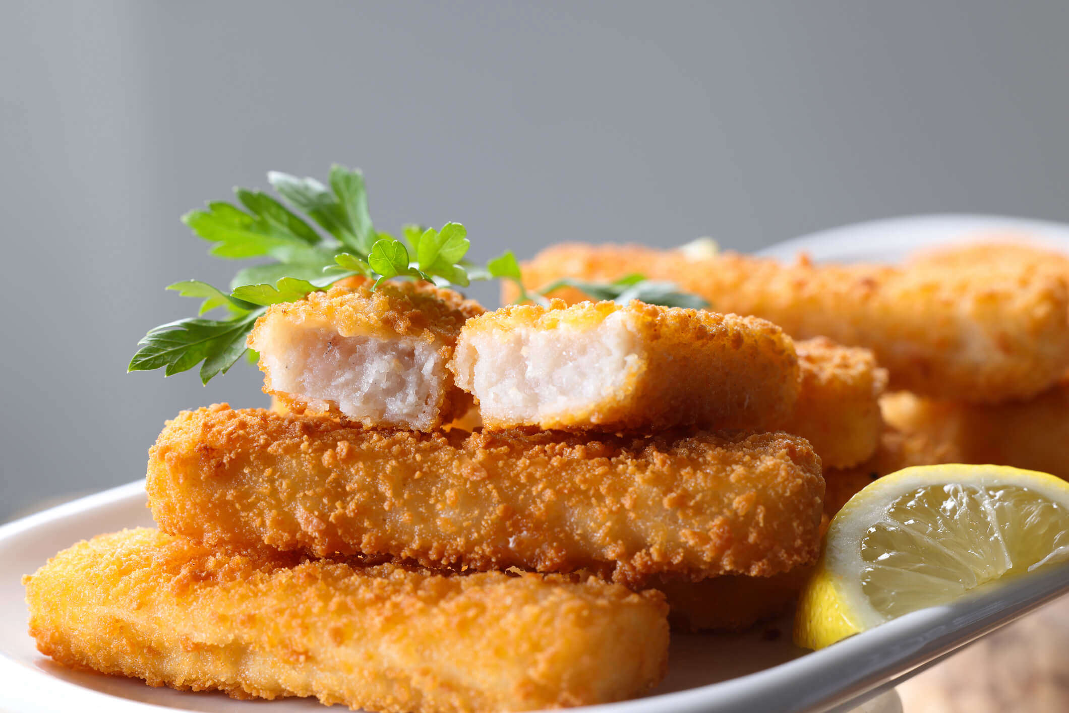 a close up of a pile of homemade fish fingers on a white plat with a slice of lemon and garnished with herbs