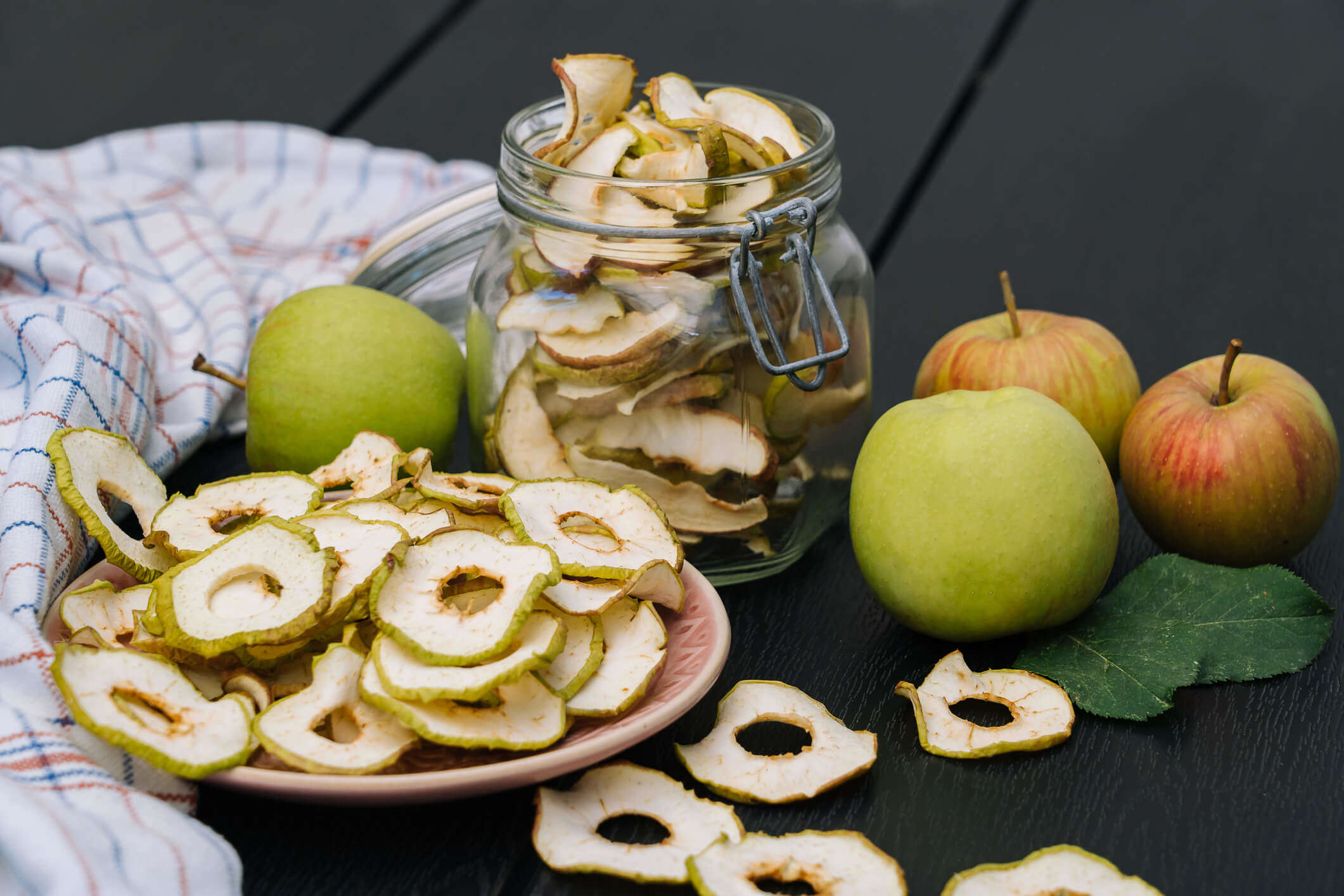 Main Image: dried apple rings, dried at home, in a jar and on plate, with fresh apples sat next to them on a table.