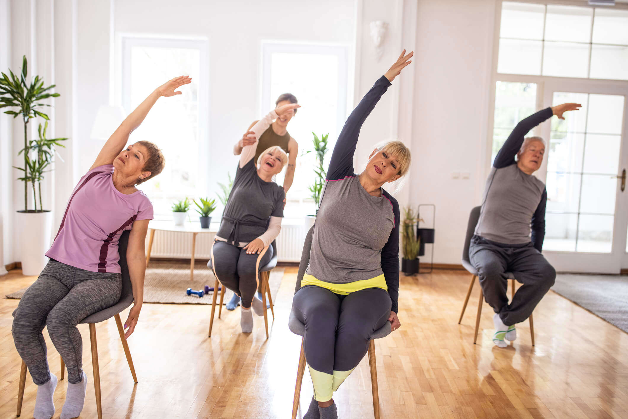 A group of older women and a man in a seated exercise class, leaning to the left with their left arm raised and stretched over their heads. With the instructor of the class helping the woman at the back with the stretch while she laughs.