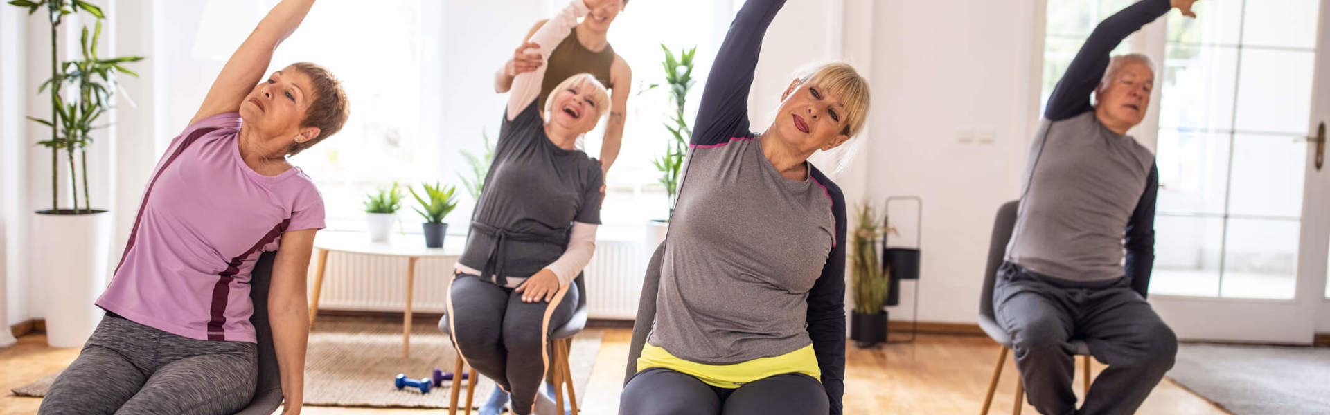 A group of older women and a man in a seated exercise class, leaning to the left with their left arm raised and stretched over their heads. With the instructor of the class helping the woman at the back with the stretch while she laughs.