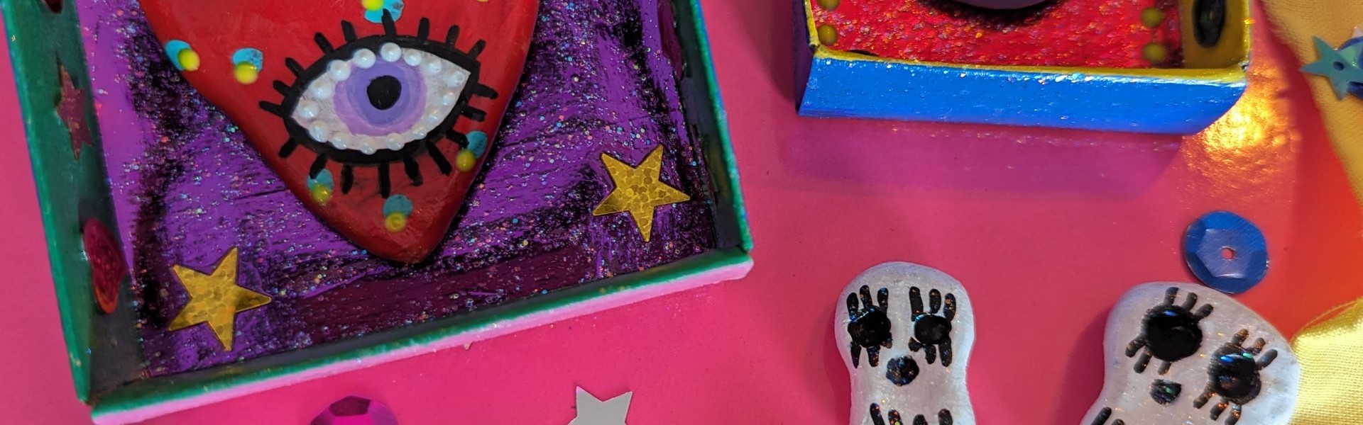 Matchbox Shrine in a variety of colours made with paper and sequins with air dry clay decorations in the shape of skulls against a pink and gold background