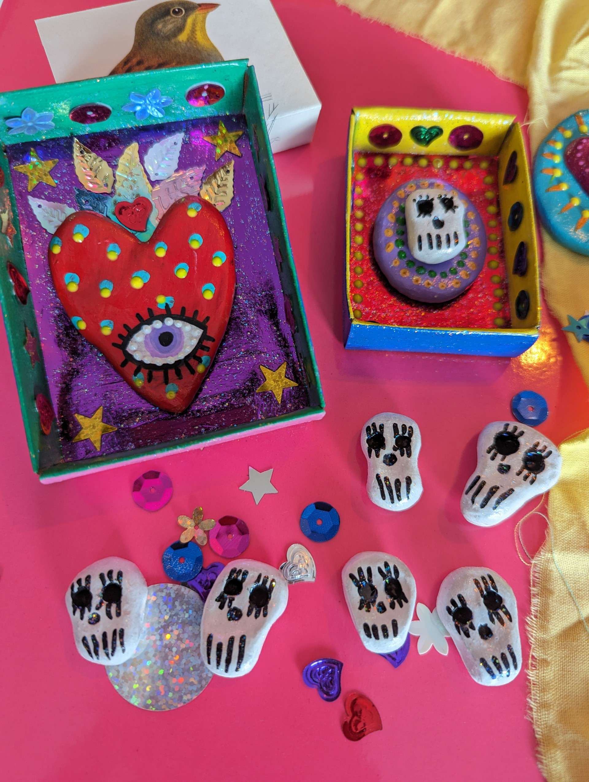 Matchbox Shrine in a variety of colours made with paper and sequins with air dry clay decorations in the shape of skulls against a pink and gold background