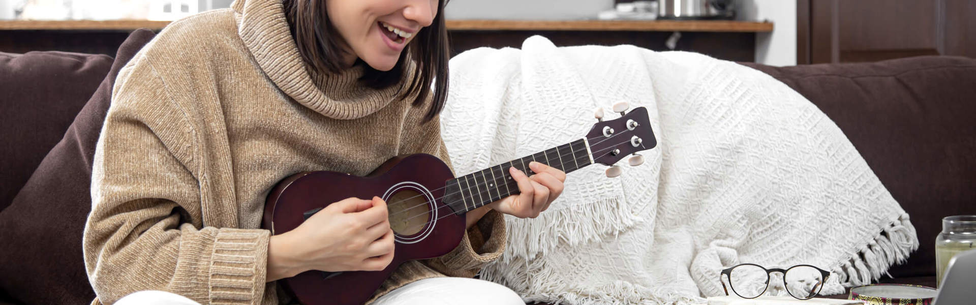 a woman sat on the sofa learning to play the ukulele from her laptop